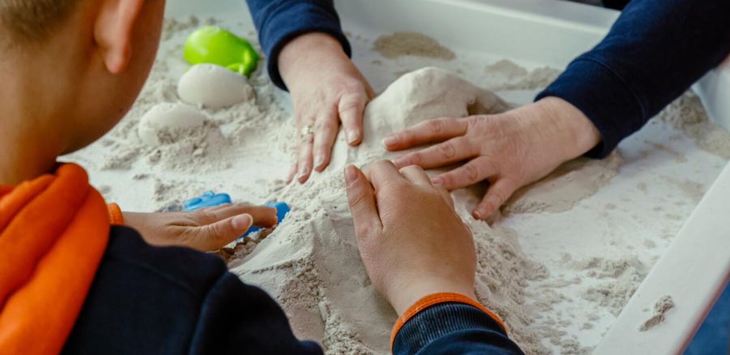 Ein Kind und eine Erzieherin formen unterschiedliche Formen in einem Sandkasten, der auf einem Tisch steht