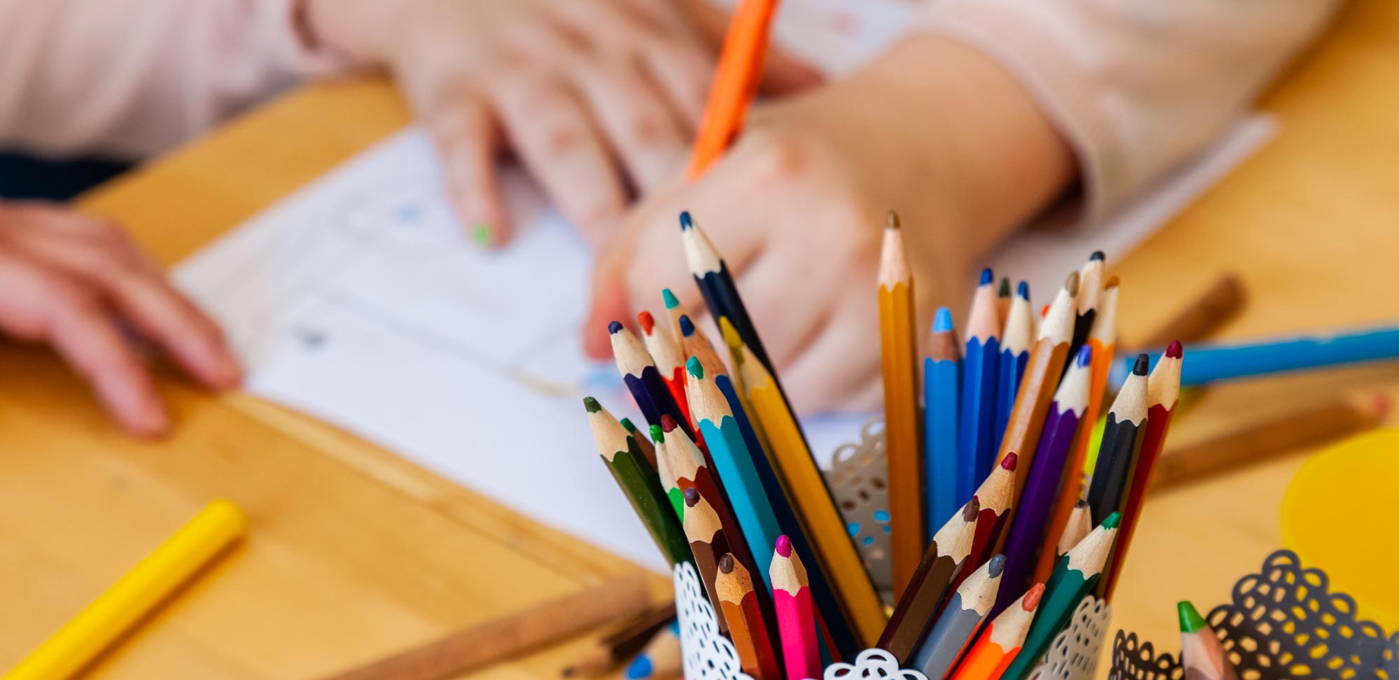 Buntstifte in einem Becher. Im Hintergrund Kinderhände beim malen zu sehen. 